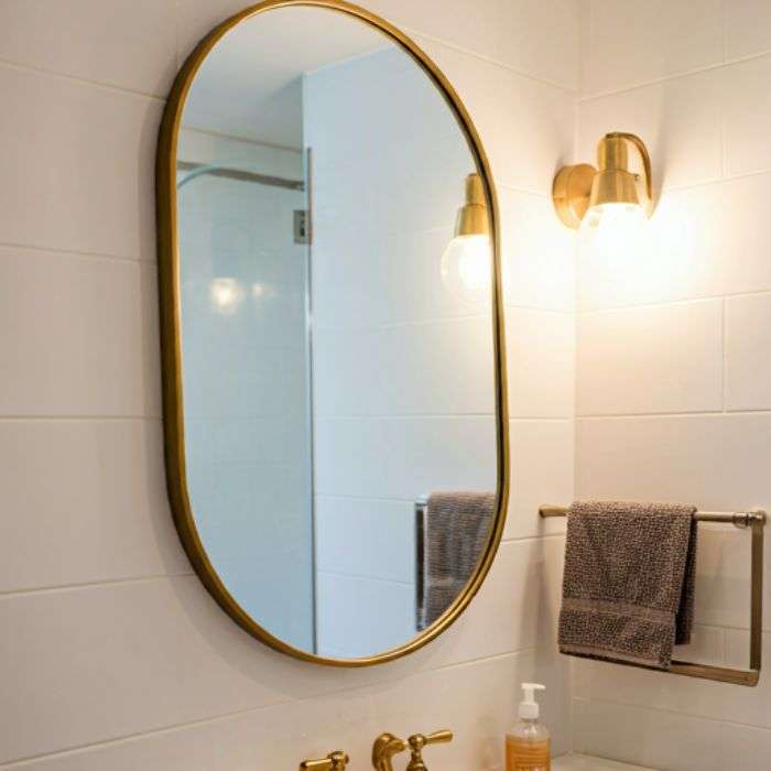 Bathroom Wall Lights: Tips for a Well-Lit Vanity and Relaxing Atmosphere A modern bathroom vanity with a white countertop and a gold-framed oval mirror. Two gold-colored wall sconces with glass shades flank the mirror, providing ample lighting.
